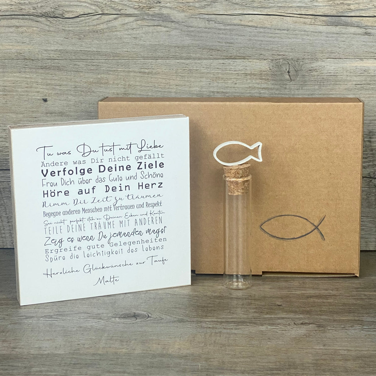 Ein liebevolles Geschenkset zur Taufe – handgefertigt und personalisierbar mit dem Namen des Kindes. Enthalten ist ein Wooden Block Art „Taufe“ (15 x 15 x 2,2 cm) mit inspirierenden Botschaften wie Tue was Du tust mit Liebe, Freu Dich über das Gute und Schöne, Hör auf Dein Herz. Dazu ein Reagenzglas mit Korken und Deko-Fisch (13 x 2,5 cm), ideal für Geldgeschenke oder Gutscheine. Verpackt in einer stilvollen Geschenkbox mit christlichen Motiven, wählbar als Fisch oder Kreuz. Nachhaltig produziert, jedes Stü