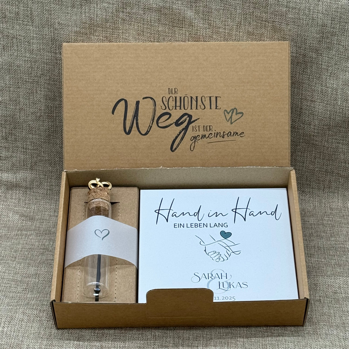 Dieses personalisierte Hochzeits-Geschenkset ist eine liebevolle Erinnerung für das Brautpaar. Es enthält einen dekorativen Holzblock in zwei romantischen Varianten – „Hand in Hand“ oder „Seite an Seite“ – personalisierbar mit Namen und Hochzeitsdatum. Dazu gehört ein Reagenzglas mit Korken und kleinen Trauringen aus Pappelholz, ideal für Geldgeschenke oder kleine Botschaften. Alles ist in einer stilvollen Geschenkbox verpackt.