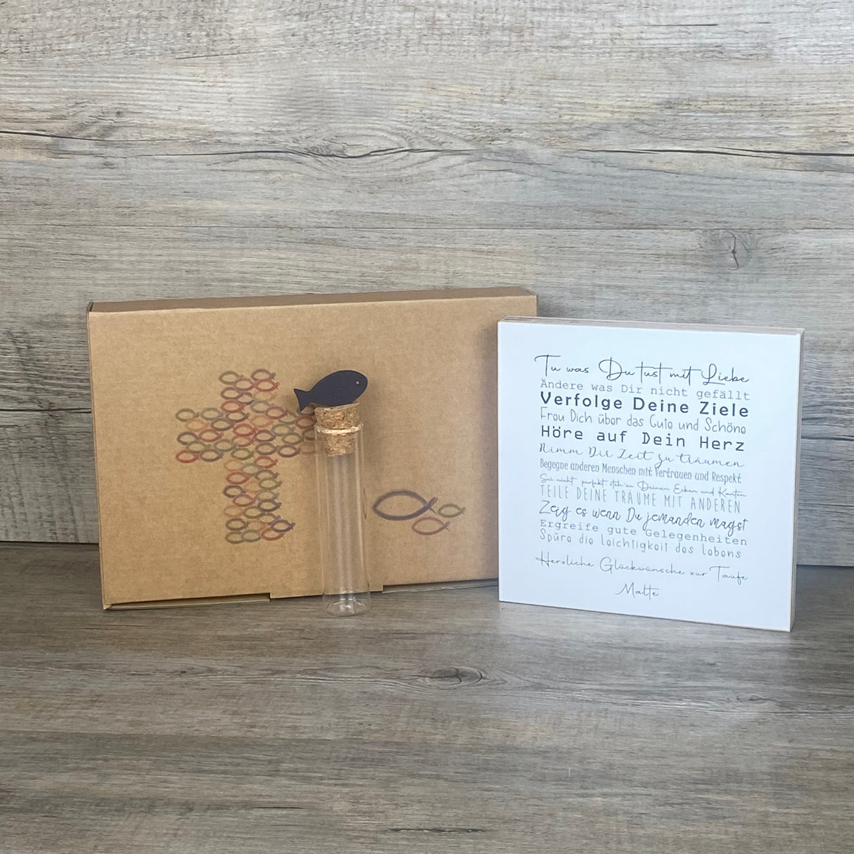 Ein liebevolles Geschenkset zur Taufe – handgefertigt und personalisierbar mit dem Namen des Kindes. Enthalten ist ein Wooden Block Art „Taufe“ (15 x 15 x 2,2 cm) mit inspirierenden Botschaften wie Tue was Du tust mit Liebe, Freu Dich über das Gute und Schöne, Hör auf Dein Herz. Dazu ein Reagenzglas mit Korken und Deko-Fisch (13 x 2,5 cm), ideal für Geldgeschenke oder Gutscheine. Verpackt in einer stilvollen Geschenkbox mit christlichen Motiven, wählbar als Fisch oder Kreuz. Nachhaltig produziert, jedes Stü