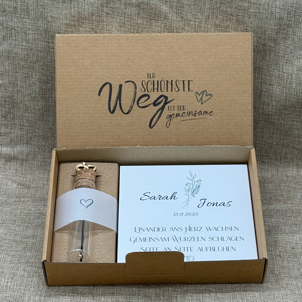 Personalisiertes Hochzeits-Dieses personalisierte Hochzeits-Geschenkset ist eine liebevolle Erinnerung für das Brautpaar. Es enthält einen dekorativen Holzblock in zwei romantischen Varianten – „Hand in Hand“ oder „Seite an Seite“ – personalisierbar mit Namen und Hochzeitsdatum. Dazu gehört ein Reagenzglas mit Korken und kleinen Trauringen aus Pappelholz, ideal für Geldgeschenke oder kleine Botschaften. Alles ist in einer stilvollen Geschenkbox verpackt.