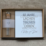 Dieses liebevolle Geschenkset zum 60. Hochzeitstag, der Diamantenen Hochzeit, besteht aus einem handgefertigten Holzblock mit inspirierenden Worten wie Lachen, Lieben und Wachsen. Auf Wunsch kann der Block mit Namen und Datum personalisiert werden. Dazu gibt es ein dekoratives Reagenzglas mit Holz-Zahl '60' für Geldgeschenke oder Gutscheine. Alles ist stilvoll verpackt in einer Geschenkbox mit Aufdruck „Der schönste Weg ist der Gemeinsame“.
