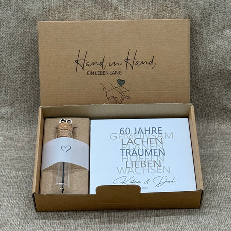 Dieses liebevolle Geschenkset zum 60. Hochzeitstag, der Diamantenen Hochzeit, besteht aus einem handgefertigten Holzblock mit inspirierenden Worten wie Lachen, Lieben und Wachsen. Auf Wunsch kann der Block mit Namen und Datum personalisiert werden. Dazu gibt es ein dekoratives Reagenzglas mit Holz-Zahl '60' für Geldgeschenke oder Gutscheine. Alles ist stilvoll verpackt in einer Geschenkbox mit Aufdruck „Der schönste Weg ist der Gemeinsame“.
